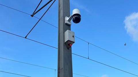 An einem Mast der Straßenbahn-Oberleitung hängt eine Kamera. Sie ist rund, darunter ein grauer Kasten mit einer kleinen, rot leuchtenden Lampe.