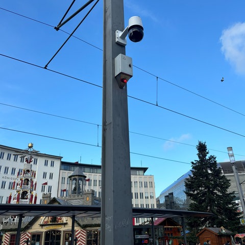 An einem Mast der Straßenbahn-Oberleitung hängt eine Kamera. Sie ist rund, darunter ein grauer Kasten mit einer kleinen, rot leuchtenden Lampe.