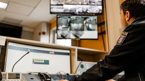 Ein Polizeibeamter sitzt in Uniform vor mehreren Bildschirmen. Darauf zu sehen sind Live-Bilder von einer Videoüberwachung im öffentlichen Raum.