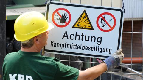 Ein Mann mit Helm hängt ein Schild mit der Aufschrift "Achtung Kampfmittelbeseitigung" an einen Zaun