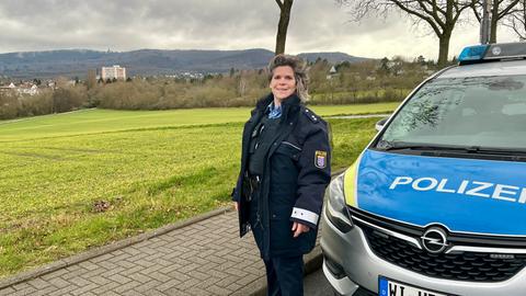 Eine Polizistin steht vor ihrem Streifenwagen. Sie hat mittellange Haar und Locken. Hinter ihr sieht man eine Stadt im Tal liegen.