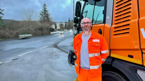 Ein Mann mit Bart steht in orangener Arbeitskleidung vor einer Kehrmaschine. Im Hintergrund fährt ein Auto eine Straße herunter.