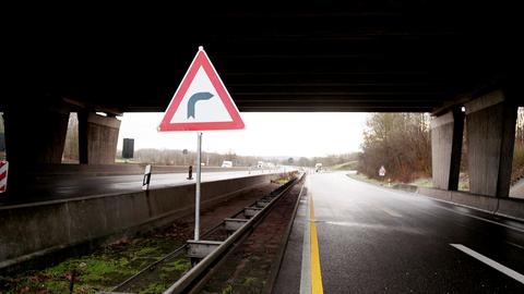 Foto von einer Kurve, die hinter einer Brücke liegt (Autobahnabfahrt/-auffahrt). Im Bildvordergrund recht groß ein Straßenschild mit einem Kurvenpiktogramm.