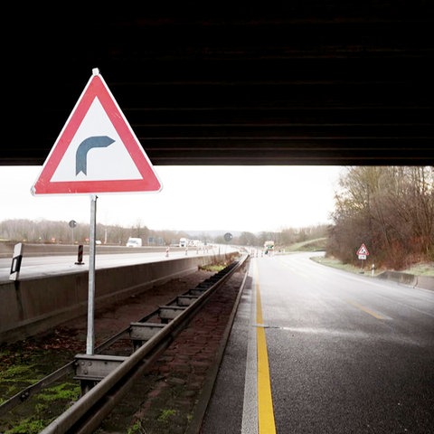 Foto von einer Kurve, die hinter einer Brücke liegt (Autobahnabfahrt/-auffahrt). Im Bildvordergrund recht groß ein Straßenschild mit einem Kurvenpiktogramm.