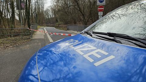 Ein Polizeiwagen steht auf einer Straße. Dahinter sieht man eine Absperrung mit einem rot-weißen Flatterband und Büsche.