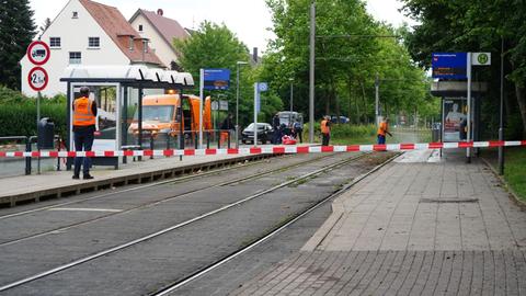 Das Bild zeigt ein rot-weißes Polizeiabsperrband an einer Straßenbahnhaltestelle. Im Hintergrund sind drei mit orangefarbenen Warnwesten bekleidete Personen zu sehen, die den Unfallort reinigen.