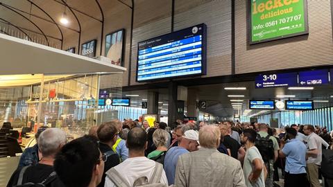 Reisende stehen am Bahnhof Wilhelmshöhe vor einer Anzeigetafel. 