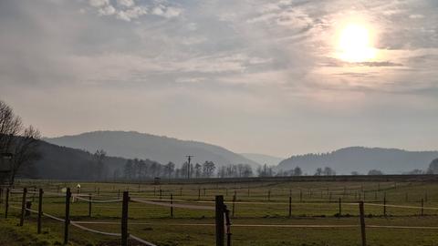 Koppeln mit Zäunen, im Hintergrund Berge, Sonne scheint durch dichte Wolken, es ist diesig.