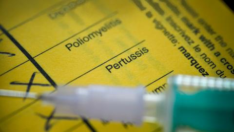 Eine Spritze liegt auf einem gelben Impfausweis. Zu lesen ist "Pertussis" und "Poliomyelitis", darunter Felder, in denen einzelne Impfungen mit Kugelschreiber angekreuzt sind.