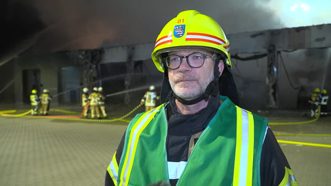 Bezirksbrandinspektor Lars Schäfer in Feuerwehrausrüstung vor der brennenden Halle