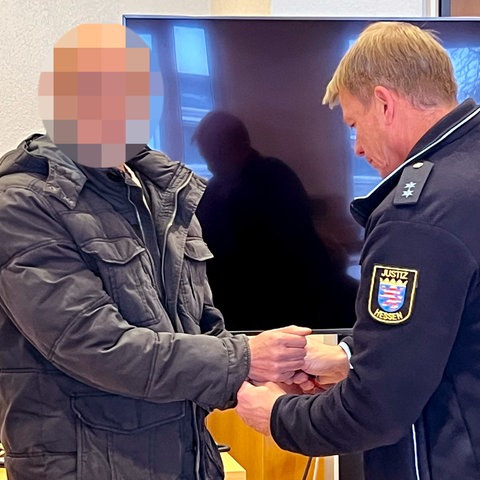 Ein Mann mit verpixeltem Gesicht hält die Hände einem Justizbeamten in Uniform entgegen, der Handschellen abnimmt.