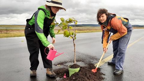 Zwei Aktivistinnen der Letzten Generation pflanzen mit Schaufel und Erde einen kleinen Baum auf den Rollweg am Airport Kassel. 