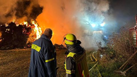 Das Bild zeigt zwei Feuerwehrmänner von hinten, die auf brennende Strohballen blicken.