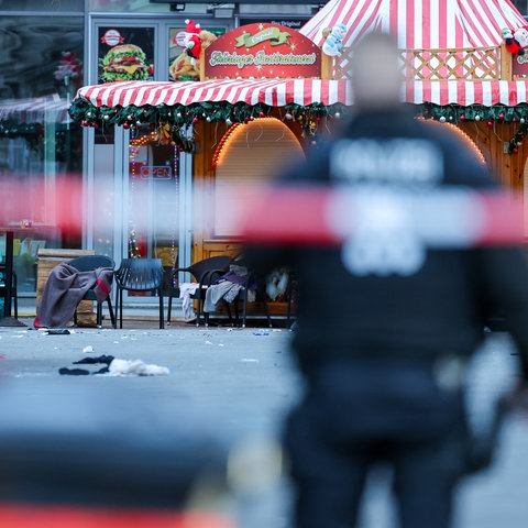 Das Bild zeigt einen leeren Stand auf dem Weihnachtsmarkt in Halle am Tag nach dem Anschlag. Im Vordergrund ist unscharf ein rot-weißes Absperrband mit der Aufschrift "Polizei" abgebildet, davor ebenfalls unscharf ein Polizist in blauer Uniform zu sehen.