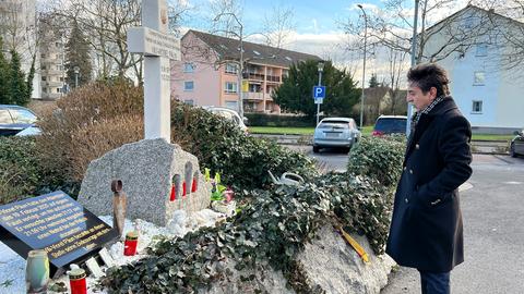 Vater Niculescu Păun in schwarzem Mantel steht vor einem großen Gedenkstein seines getöteteten Sohnes, daneben Kerzen. Im Hintergrund ein Parkplatz und eine Wohnsiedlung.