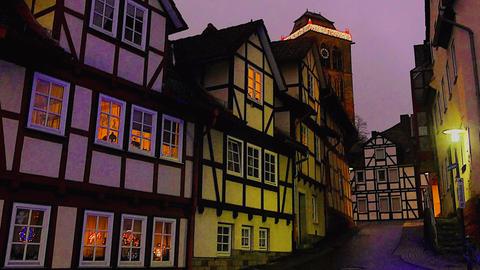 Fachwerkhäuser an einer schmalen Gasse im abendlichen Dämmerlicht. In einigen Fenstern brennt Licht, im Hintergrund ist ein eckiger Turm zu sehen, dessen Spitze mit Weihnachtslichtern geschmückt ist.