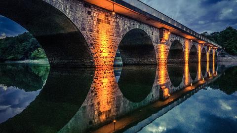 Bild einer Brücke mit angestrahlten Brückenpfeilern über einen See am Abend