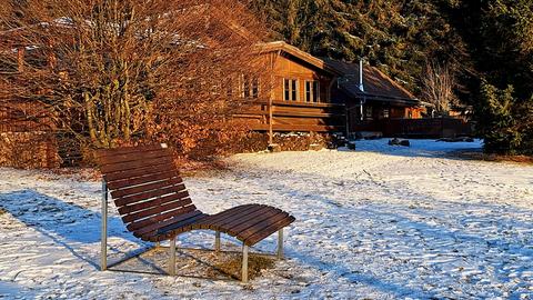 Geschwungene Sitzbank auf schneebeckter Wiese vor Holzfassade von Haus in der Sonne