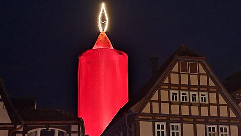 Ein Fachwerkhaus bei Nacht. Dahinter ein runder Turm, der in ein rotes Tuch eingehüllt ist und mit einer Spitze in Form einer gelben Kerzenflamme versehen ist