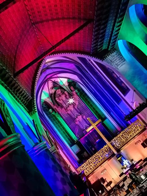 Bunt (rot, blau, lila, grün) angestrahltes Innere der Universitätskirche Marburg, Blick auf den Altar, die Orgel und ein großes goldenes Kreuz; schräg gehaltene Kamera