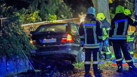 Feuerwehrleute stehen im Dunkeln vor einem Auto, das gegen einen Baum gefahren ist