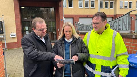 Eine Frau und zwei Männer stehen vor einem Behördenbau. Sie schauen gemeinsam auf einen Plan.