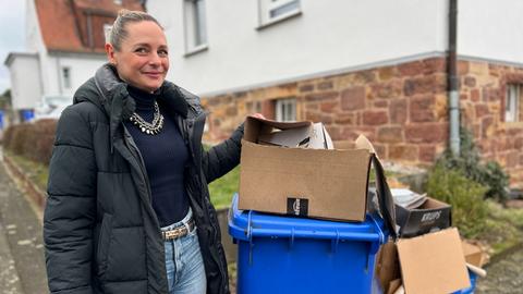 Eine Frau steht neben einer blauen Mülltonne. Ein Karton steht auf dem Deckel, darin weiteres Altpapier.