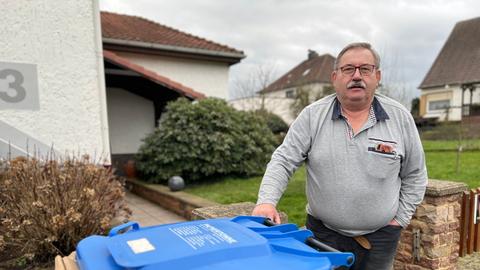 Ein Mann steht vor seinem Haus an einer blauen Mülltonne voller Altpapier.