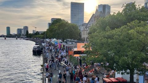 Am Mainufer in Frankfurt stehen Essensstände, dazwischen laufen Besucherinnen und Besucher eines Festes.