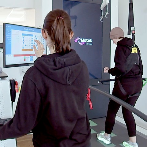 Zwei Frauen an einem Reha-Gerät. Eine Frau steht auf dem Gerät, eine Sturzsicherung von oben hält sie in einem Gurt fest. Daneben eine Therapeutin am Computer. 