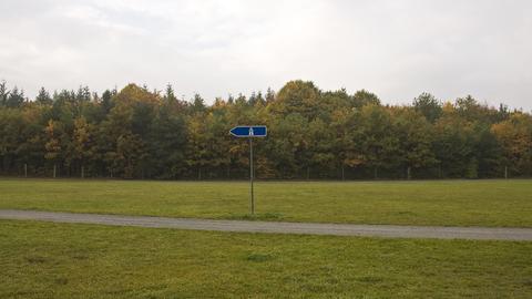 Ein blaues Autobahnschild steht auf einer weitläufigen Grasfläche, durch die ein Weg verläuft. Im Hintergrund grenzt ein Wald an.  