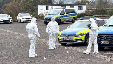 Drei Polizeiautos stehen auf einem Parkplatz. Daneben drei Menschen in weißen Ganzkörperanzügen und Markierungen auf dem Boden.