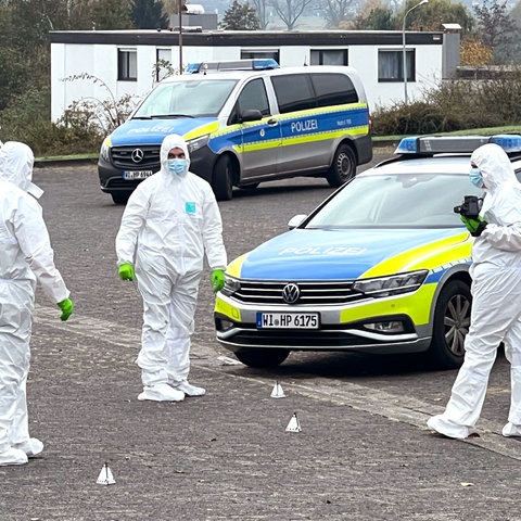 Drei Polizeiautos stehen auf einem Parkplatz. Daneben drei Menschen in weißen Ganzkörperanzügen und Markierungen auf dem Boden.