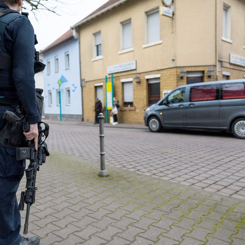 Bildvordergrund (scharf): Polizist mit Maschinengewehr steht an einer Straße. Bildhintergrund (leicht unscharf): Gebäude mit Eckkneipe, davor steht ein Leichenwagen.