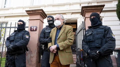 Ein Mann mit FFP2-Maske und Handschellen wird von mehreren Polizeibeamten aus einem Haus geführt. 