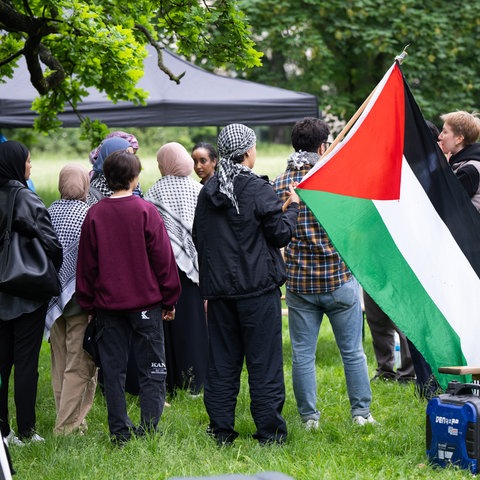 Das Bild zeigt Teilnehmende eines pro-palästinensischen Protestcamps auf dem Campus der Goethe Universität. Sie stehen unter einem auf einer Wiese aufgebauten Pavillon, tragen Palästinensertücher und schwenken die palästinensische Fahne.