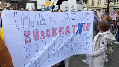 Eine Frau hält bei einer Demonstration ein weißes Bettlaken in der Hand. Darauf steht: "Immer mehr Bürokratie, keine Zeit für Patienten".