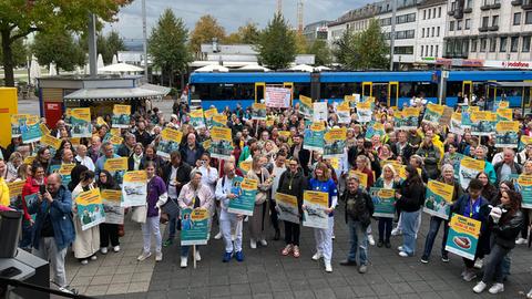 Demonstrationsteilnehmer auf einem Platz in Kassel. Sie halten Transparente mit der Aufschrift "Budgetierung abschaffen" in den Händen, im Hintergrund fährt eine Straßenbahn.