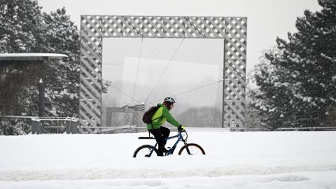 Ein Radfahrer radelt über eine verschneite Straße. Er trägt einen Helm und eine grüne Jacke. Im Hintergrund sind verschneite Bäume und ein großes Kunstwerk wie ein Rahmen aus Metall.