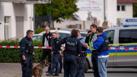 Polizisten stehen in einem abgesperrten Bereich auf einer Straße, ein Spürhund steht bei ihnen. 