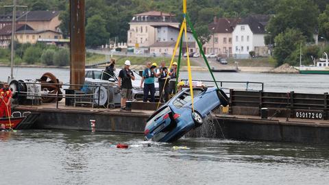 Ein Kleinwagen wird mit gelben Seilen an einem Kran hängend aus einem Fluss gezogen. Daneben Menschen, die auf einer Plattform stehen.