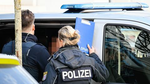 Eine Polizistin und ein Polizist von hinten vor einer offenen seitlichen Polizei-Transportertür stehend, dazwischen guckt ein verpixelter Kopf hervor. 