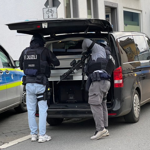 Hinter einem schwarzen Polizeiauto, am Kofferraum, stehen zwei Polizisten, einer mit Waffe.