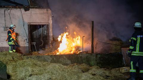 Brand in und an Scheune in der Nacht. Neben der Scheune stehen zwei Feuerwehrleute mit Schläuchen.