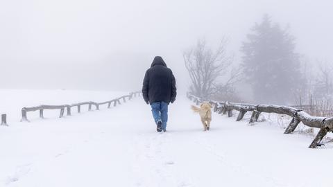 Ein Mann in Winterkleidung geht mit seinem Hund auf einem verschneiten Wanderweg.