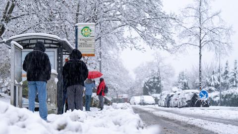 Menschen stehen im Schnee an einer Bushaltestelle in Wiesbaden.