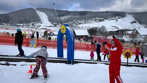 Ein paar Kinder auf Skiern, eine Frau im roten Schneeanzug gibt jemandem ein Handzeichen.