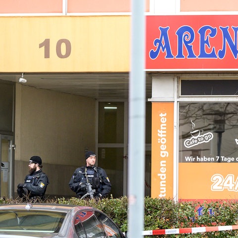 Zwei Polizisten stehen in einem Hauseingang, der mit rot-weißem Flatterband abgesperrt ist.