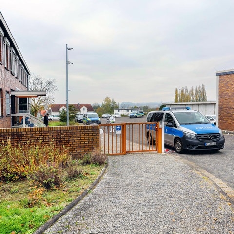 Das Bild zeigt die Polizeistation in Schwalmstadt. Zu sehen ist ein Gebäude aus rotem Backstein mit einem rostroten Zaun davor. Die Zufahrt ist durch ein Polizeiauto blockiert. Im Hintergrund sind Ermittler der Spurensicherung zu sehen.