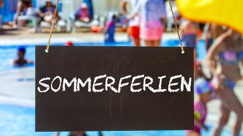  Ein Schild mit der Aufschrift "SOMMERFERIEN" vor Sommerurlaubern im Freibad.